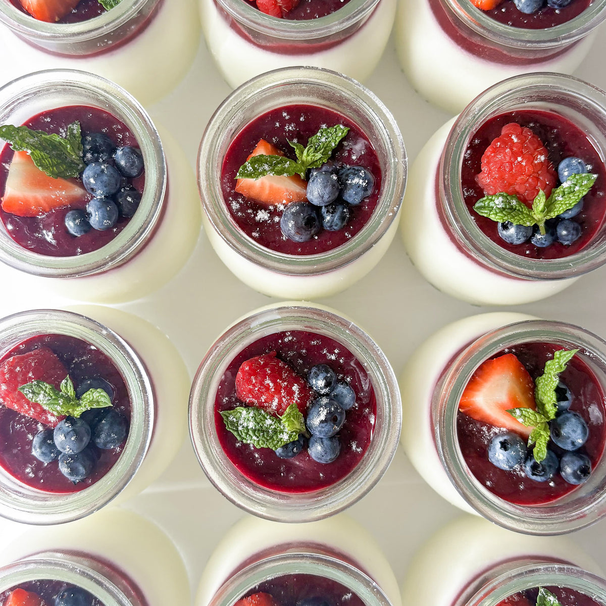 Overhead view of multiple small glass jars filled with white panna cotta, topped with dark berry coulis, fresh strawberries, blueberries, and mint leaves, lightly dusted with powdered sugar and arranged in a neat grid on a white background.