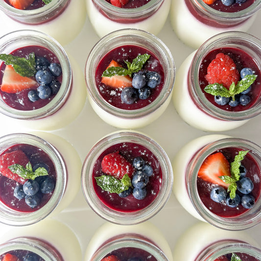 Overhead view of multiple small glass jars filled with white panna cotta, topped with dark berry coulis, fresh strawberries, blueberries, and mint leaves, lightly dusted with powdered sugar and arranged in a neat grid on a white background.