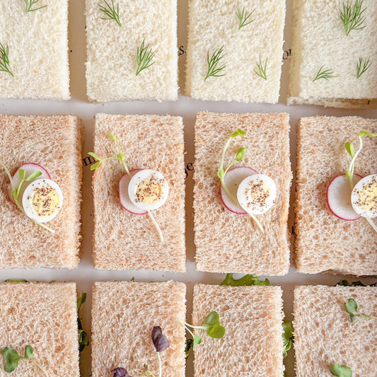 Overhead view of a grid of small tea sandwiches made with white and brown bread, topped with sliced hard-boiled eggs, radish slices, and microgreens.