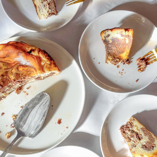 Overhead view of several white plates holding slices of a marble basque cheesecake, with crumbs and a metal pie server on a white surface.