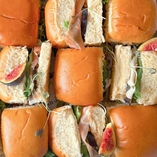Box of mini brioche sandwiches with proscuitto, figs and lettuce on a white background.