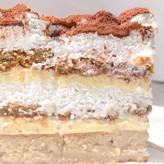 A close-up side view of a layered tiramisu cake, showing alternating layers of whipped cream, yellow pastry cream, soaked coffee lady finger cookies, and a dusting of cocoa powder on top.
