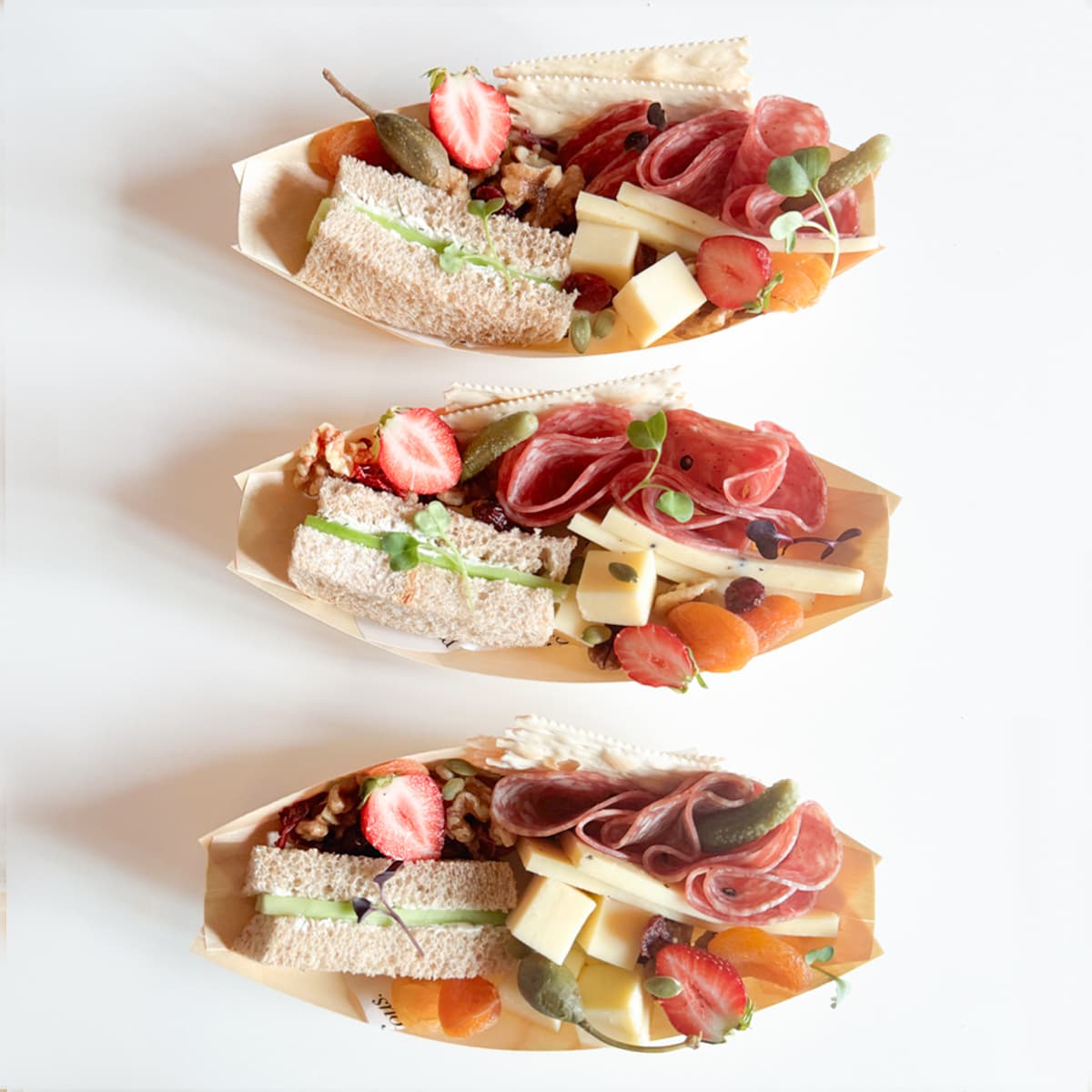 Three beautifully curated charcuterie boats arranged vertically on a clean white background, containing one cucumber tea sandwich, cheeses, cured meats, assorted nuts and seasonal fruit.