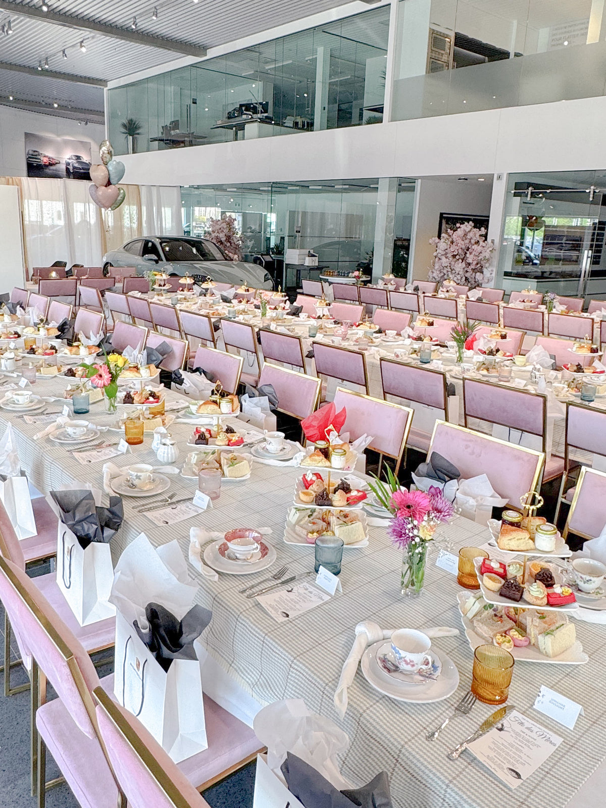 A large afternoon tea event dining hall set up with long tables covered in white tablecloths, pink chairs, and multiple tiered stands filled with sandwiches, cakes and pastries. The room has a glass upper mezzanine, and a few balloons are visible near the window.