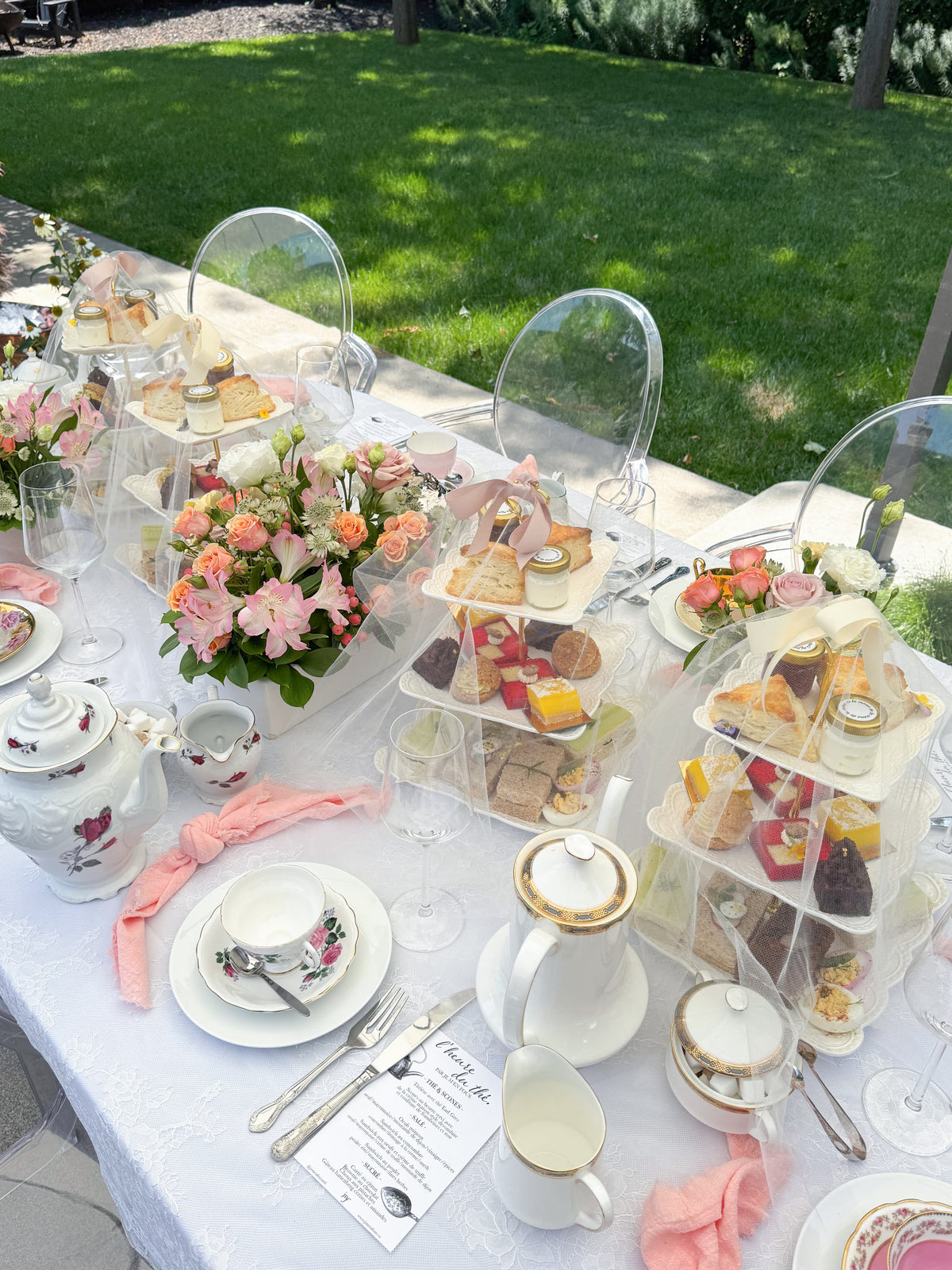 An outdoor afternoon tea table setup with white teapots, cups, saucers, floral centerpieces, and clear acrylic chairs surrounding tiered stands filled with sandwiches, cakes and pastries. A green lawn is visible in the background.