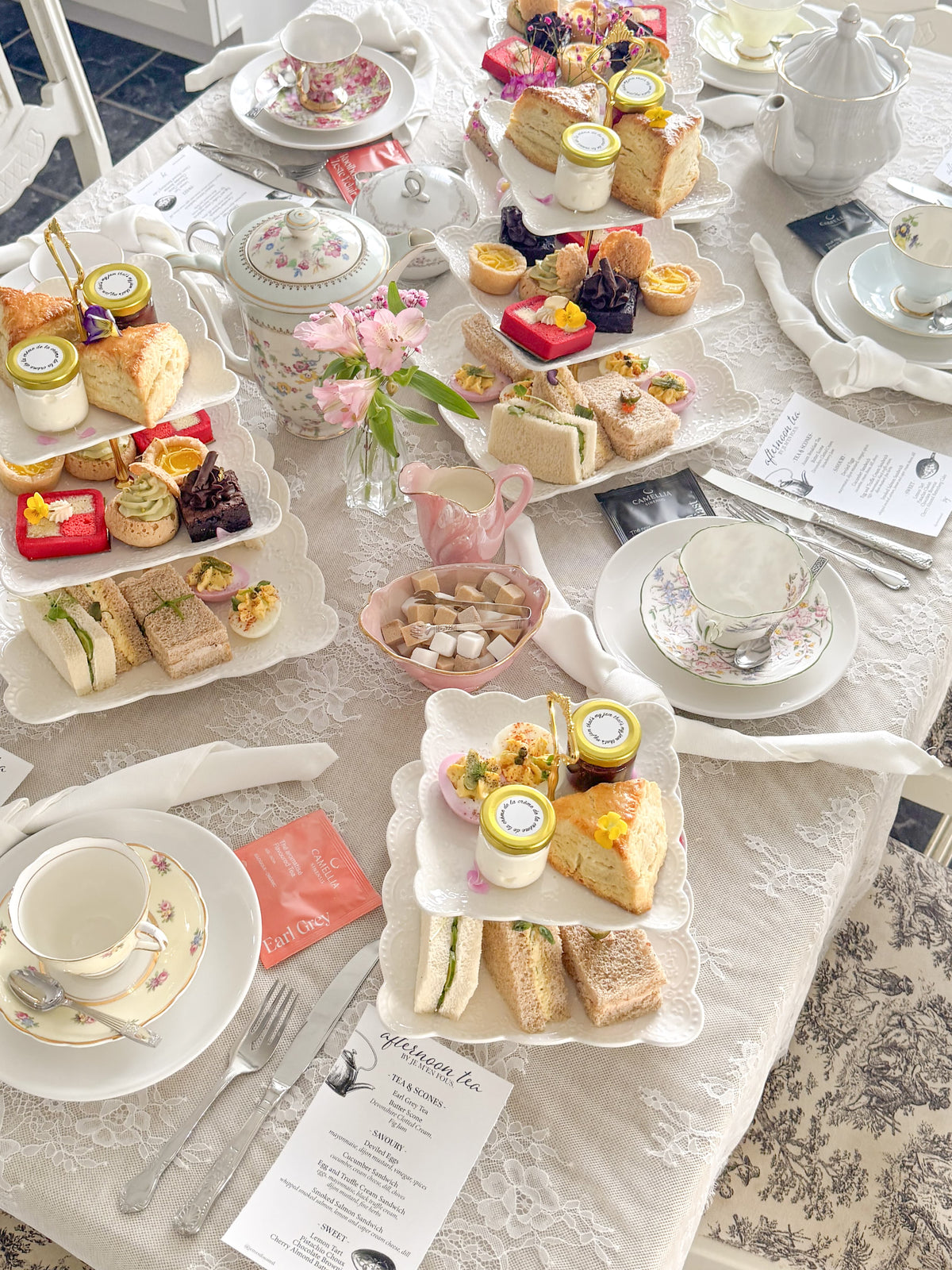 An elegant afternoon tea spread arranged on a white tablecloth, featuring multiple white ceramic serving trays, teapots, teacups, sandwiches, cakes, pastries, and small jars. A printed menu card is visible in the foreground.