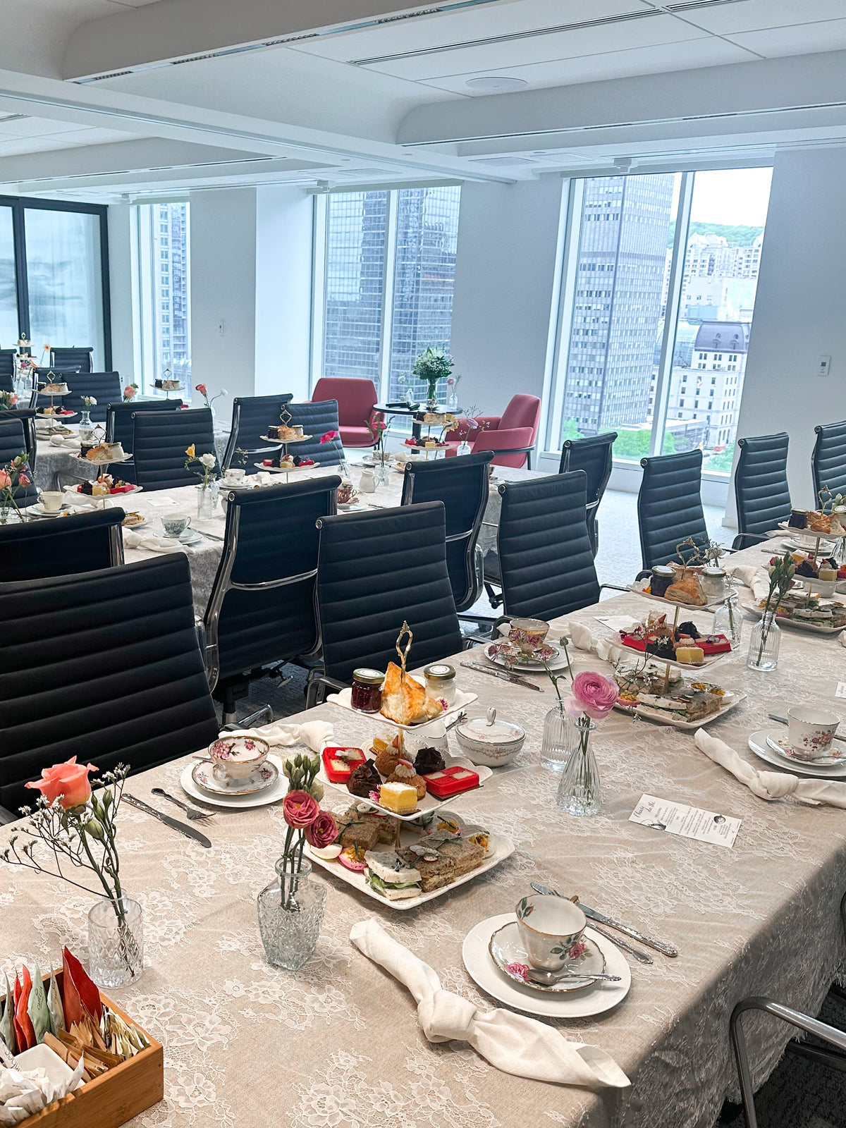 A long conference-style dining table set for tea in a bright office with large windows. Multiple place settings with teacups, saucers, plates, cutlery, and small floral arrangements are arranged along the table, surrounded by black office chairs.