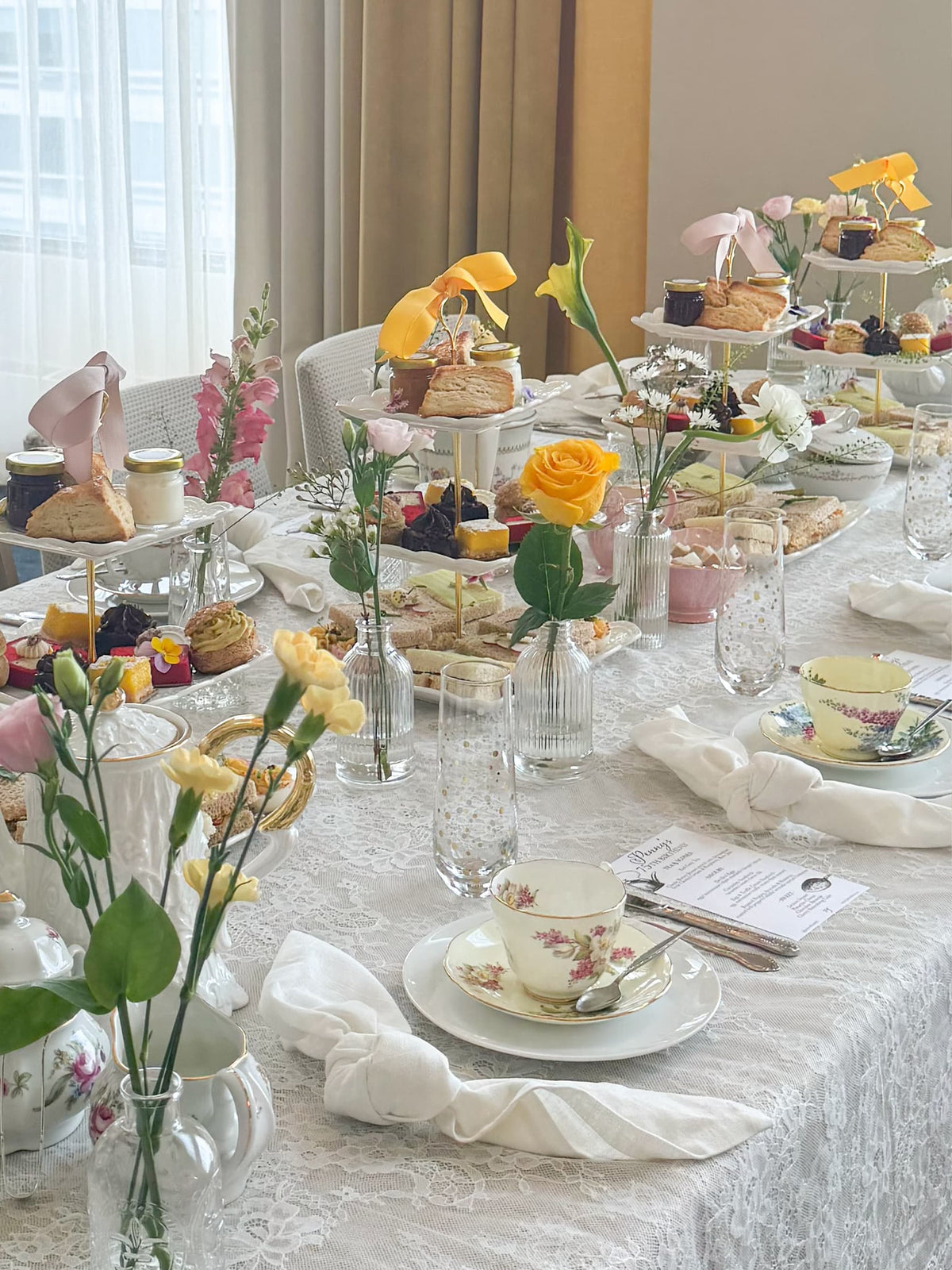 A beautifully styled tea and pastry spread on a white lace-covered table, featuring floral teacups, yellow roses, and multiple tiered stands filled with sandwiches and pastries in a bright indoor setting.