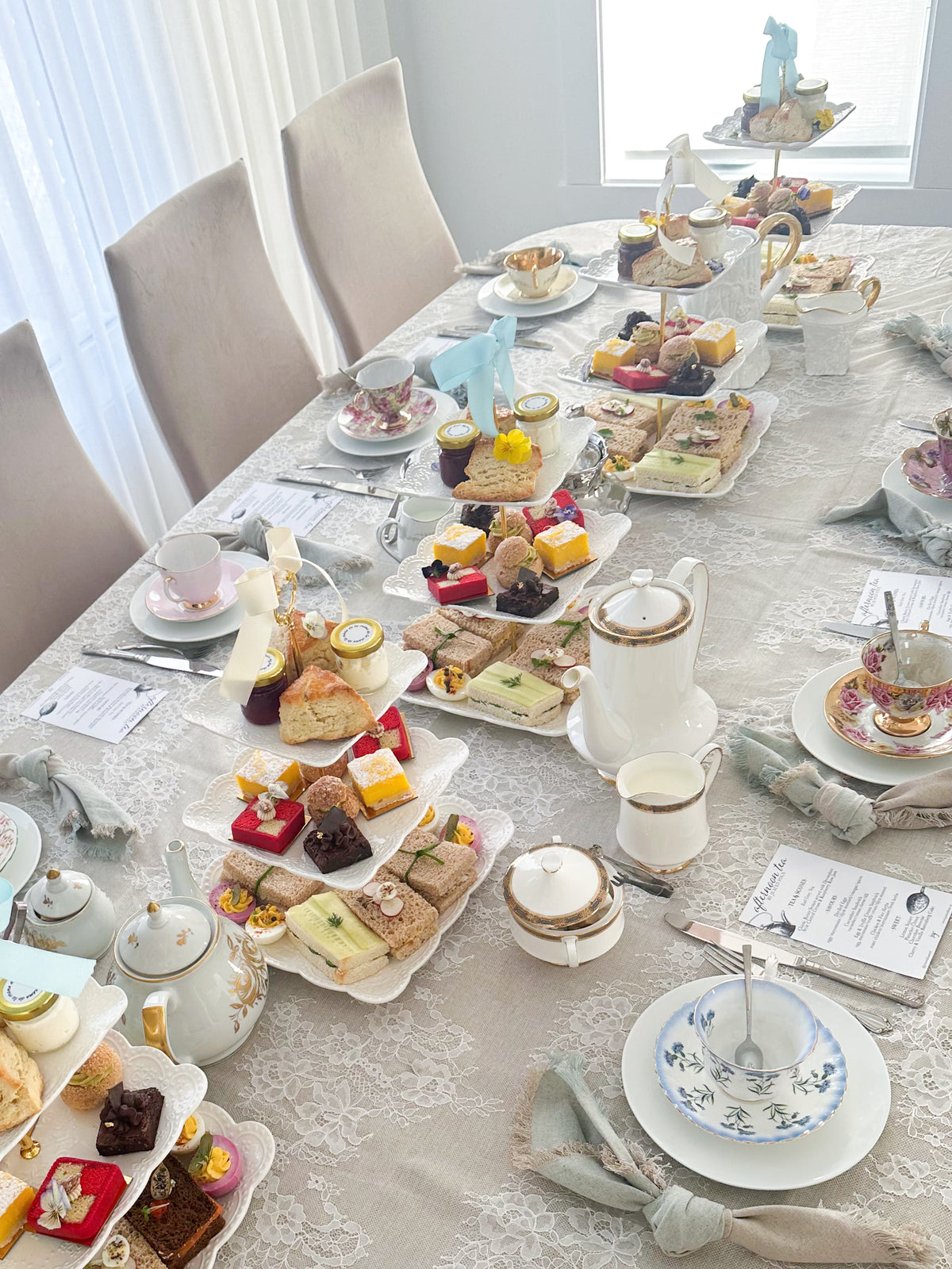 An elegant afternoon tea table setting with multiple tiers of pastries, sandwiches, cakes, teapots, teacups, and saucers arranged on a patterned white tablecloth in a bright dining room.