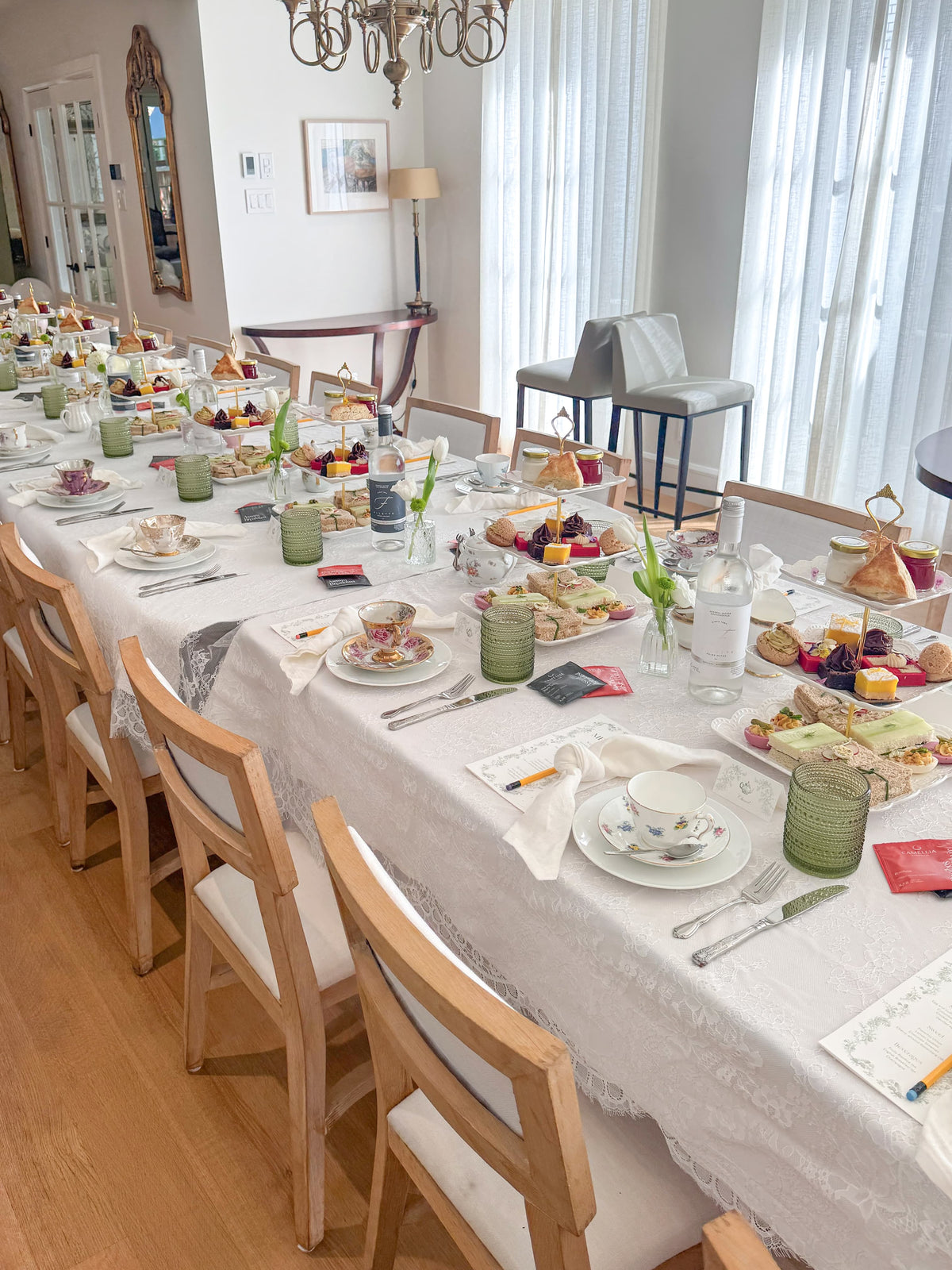A long dining table set for a tea service in a bright dining room. The table is covered with a white lace tablecloth and set with white teacups, saucers, silver cutlery, green glass cups, platters with sandwiches and mini desserts, napkins, and small green and white flowers.