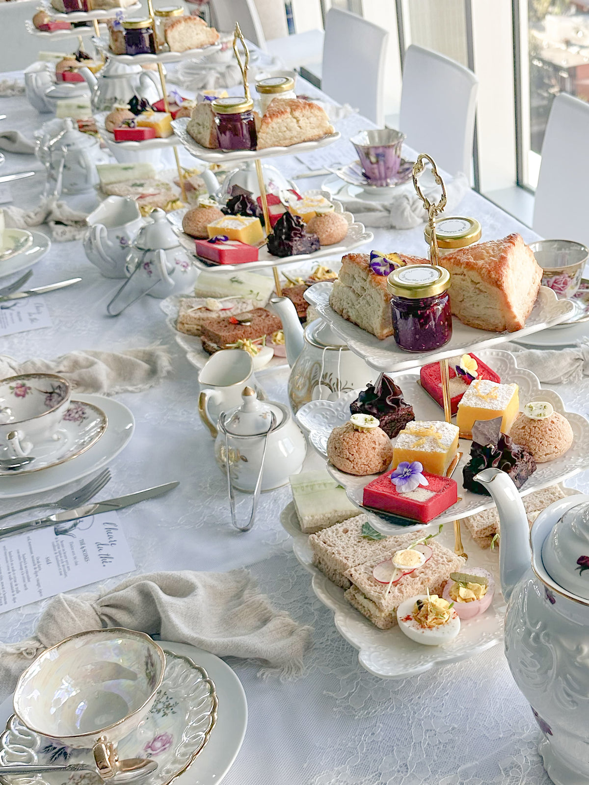 A long table set for an afternoon tea spread, with white teacups, saucers, teapots, creamers, and multiple tiered serving trays filled with sandwiches, scones, pastries, and small desserts. White chairs are visible along the table, and bright window light illuminates the scene.