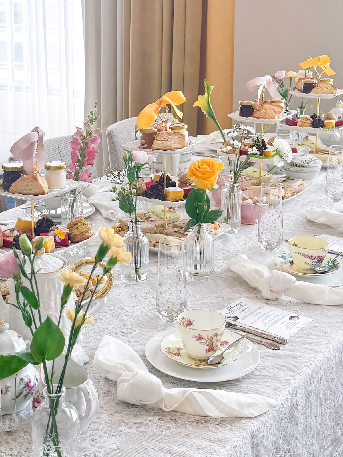 A beautifully decorated long table set for an afternoon tea event, featuring an elegant white lace tablecloth, teacups, pastries, cakes, and small floral arrangements in clear glass vases. The background shows a modern room with sheer white curtains.