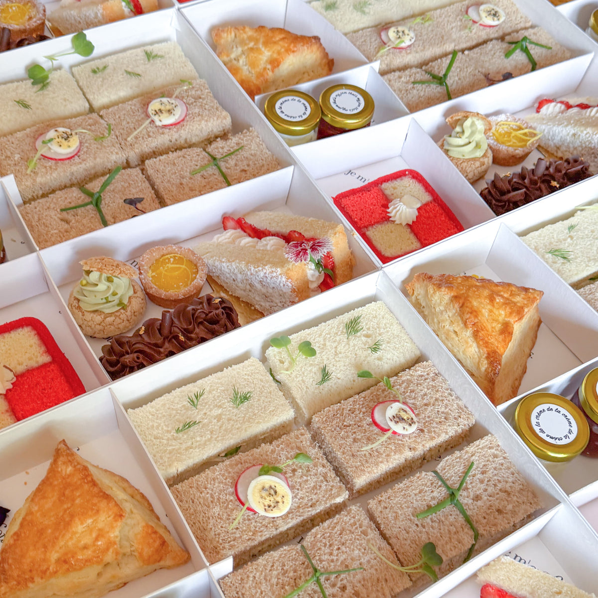 A close-up overhead view of a boxed assortment of tea sandwiches, pastries, scones and small jars arranged in white compartments. 