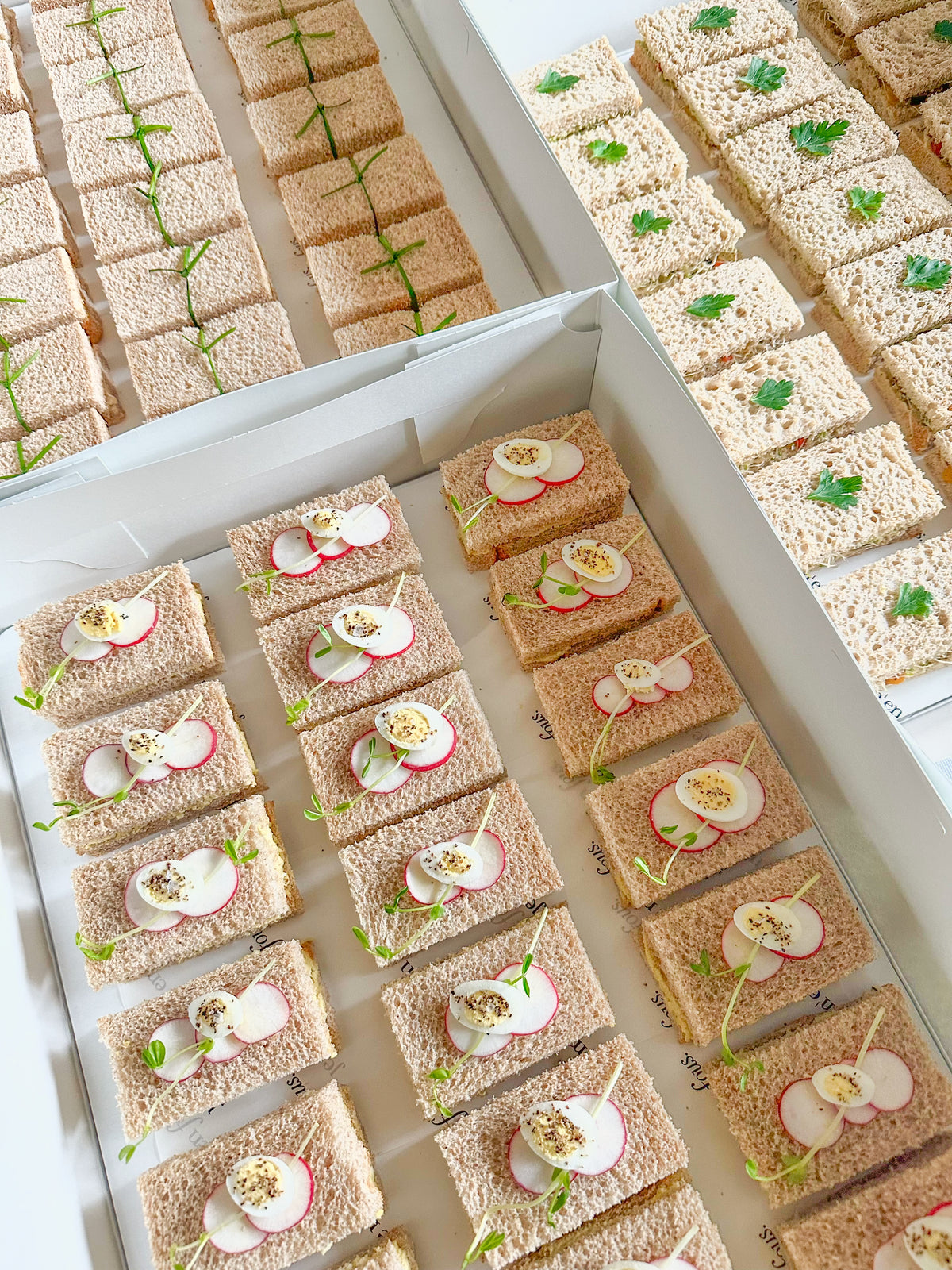 Assorted tea sandwiches, decorated with eggs, tied with a chive and decorated with microgreens. Served in white boxes.