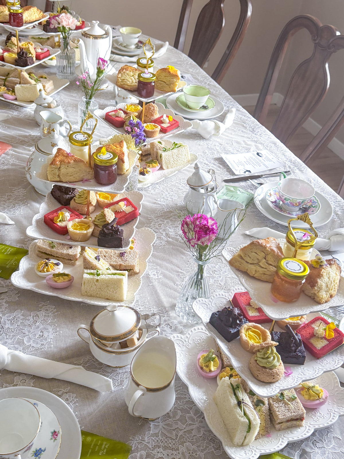 A beautifully decorated long table set for an afternoon tea event, featuring an elegant white lace tablecloth, teacups, pastries, cakes, and small floral arrangements in clear glass vases. The background shows a vintage room with brown wood chairs.