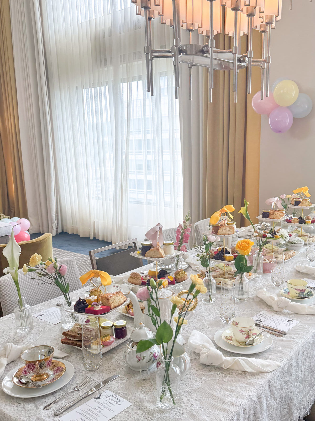 A beautifully decorated long table set for an afternoon tea event, featuring an elegant white lace tablecloth, teacups, pastries, cakes, and small floral arrangements in clear glass vases. The background shows a modern room with sheer white curtains, a multi-light chandelier, and pastel balloons.