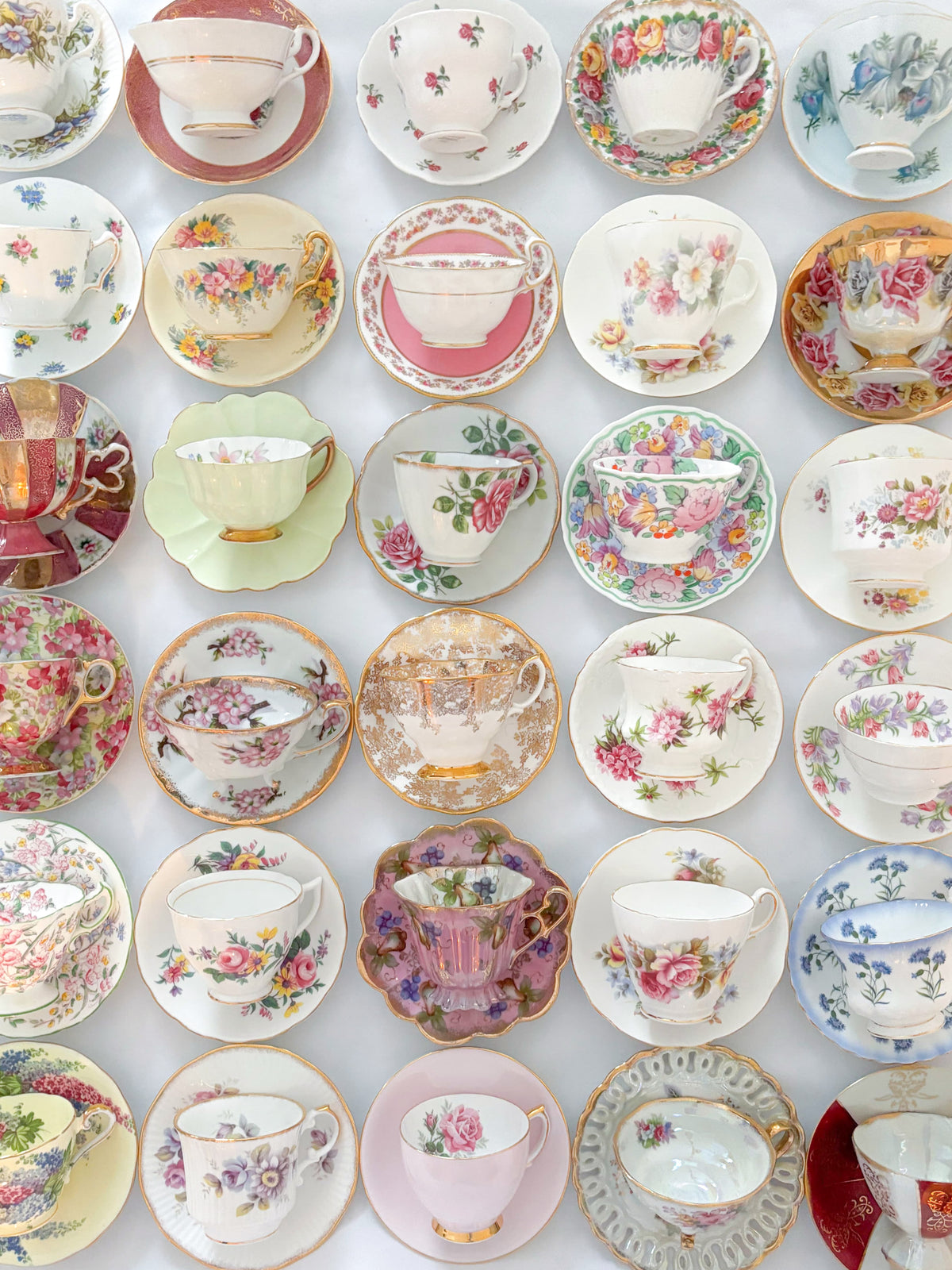 A grid arrangement of numerous vintage floral teacups and saucers in various colors, including pink, green, blue, red, and gold, displayed on a white background.