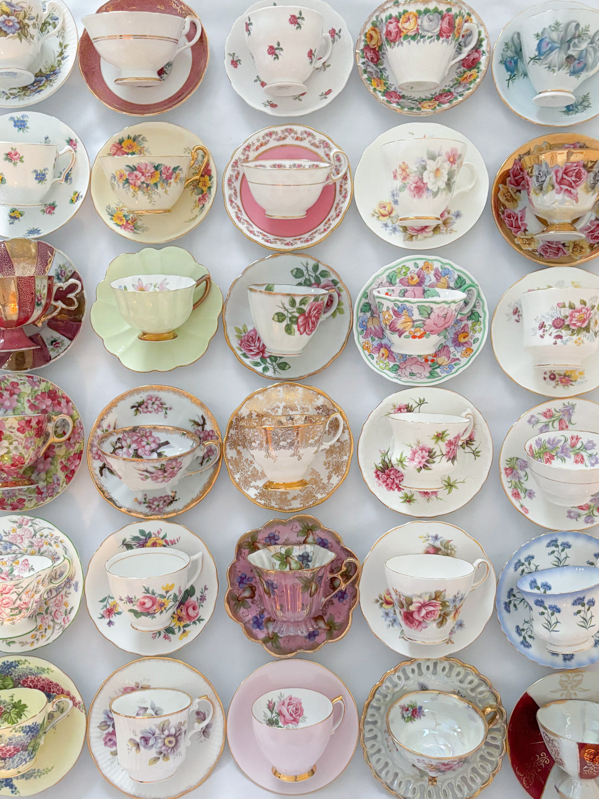 A grid arrangement of multiple vintage floral teacups and saucers in various colors, including pink, green, blue, gold, and red, displayed on a white background.