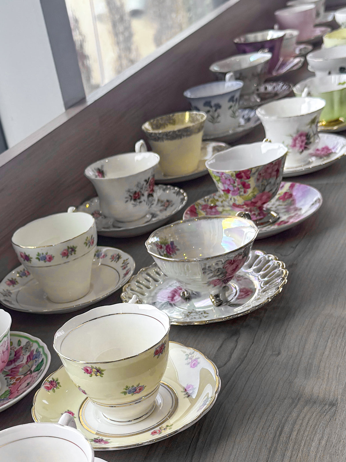 A collection of vintage floral teacups and saucers arranged diagonally on a dark wooden table, featuring delicate pink, yellow, and gold patterns.