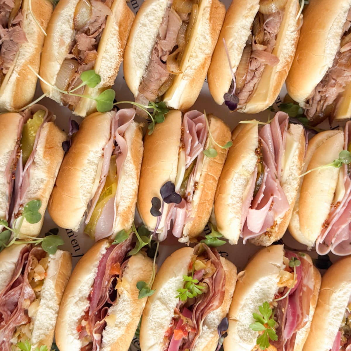 A close-up overhead view of assorted mini sub sandwiches arranged tightly together. The sandwiches are filled with sliced meats, pickles, onions and topped with microgreens.