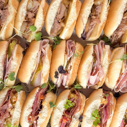 A close-up overhead view of assorted mini sub sandwiches arranged tightly together. The sandwiches are filled with sliced meats, pickles, onions and topped with microgreens.