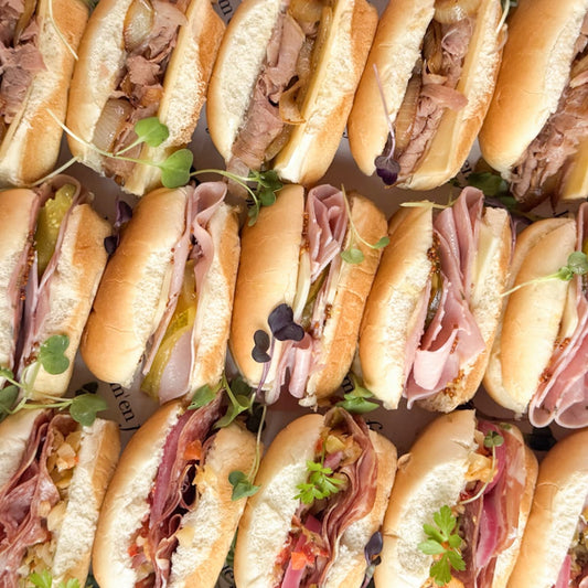 A close-up overhead view of assorted mini sub sandwiches arranged tightly together. The sandwiches are filled with sliced meats, pickles, onions and topped with microgreens.