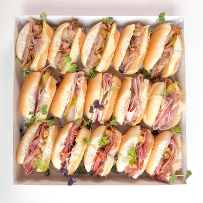 A white box filled with assorted deli meat and cheese sandwiches, garnished with microgreens, set against a clean white background.