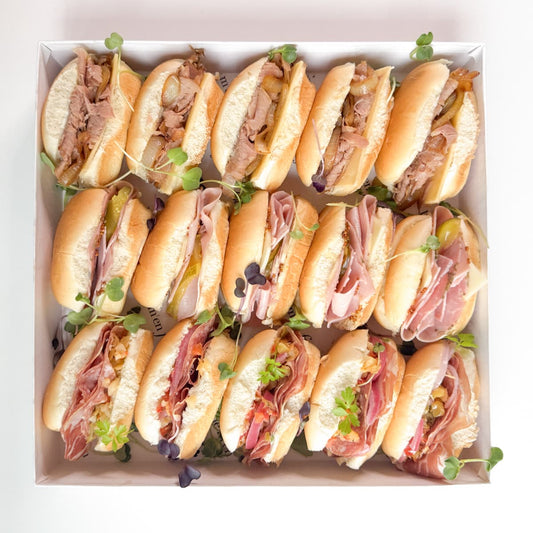 A white box filled with assorted deli meat and cheese sandwiches, garnished with microgreens, set against a clean white background.
