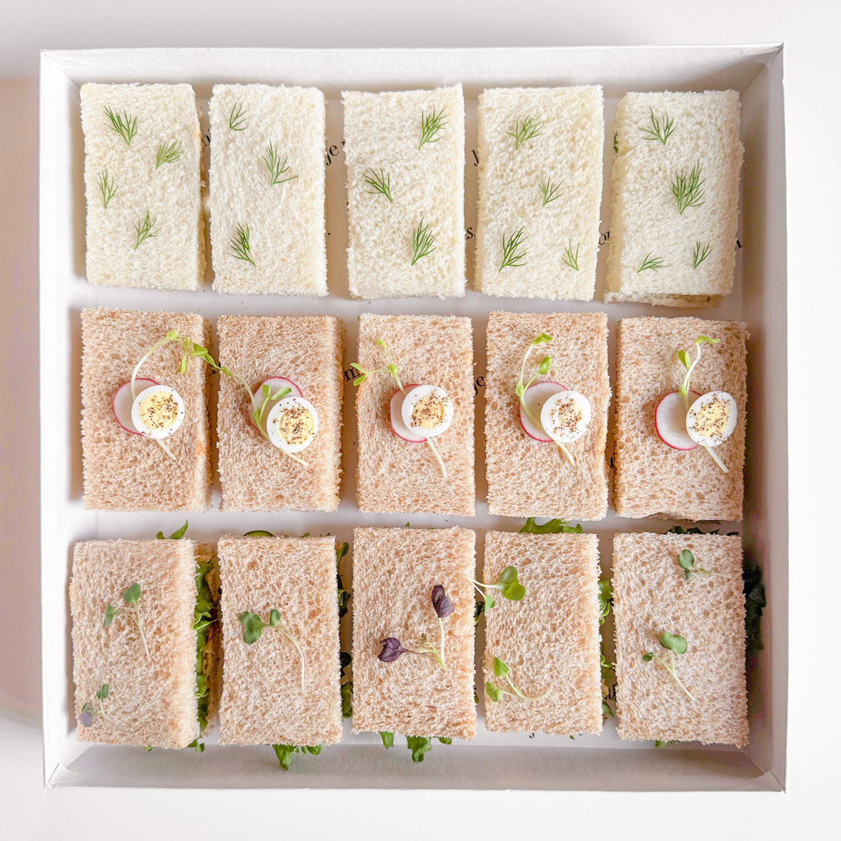 A top-down view of a white square tray holding 15 small tea sandwiches arranged in three rows. The top two rows are made of white bread with green herb garnish, while the bottom two rows are made with brown bread with small garnish accents.