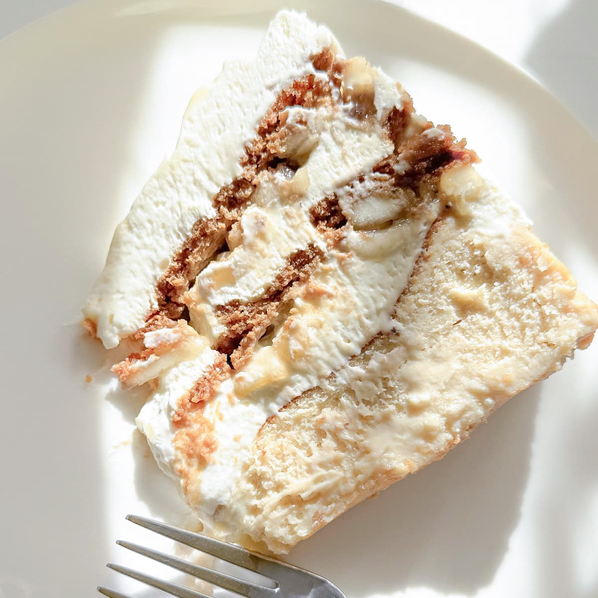 A close-up of a slice of layered banana cake on a white plate, with a fork partially visible at the bottom and strong sunlight casting shadows.