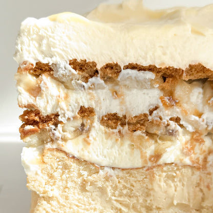 A close-up of a layered dessert slice with white whipped cream, biscoff cookie pieces, sliced bananas, and a cheesecake base.