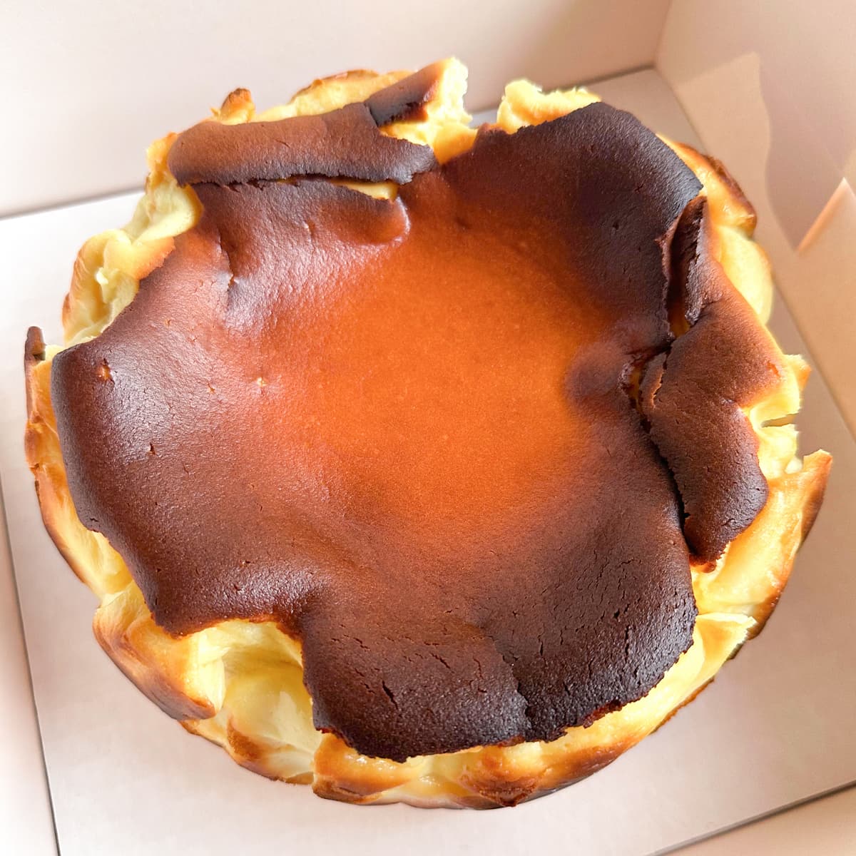A freshly baked basque cheesecake with a dark caramelized top crust, sitting inside a white cardboard box.