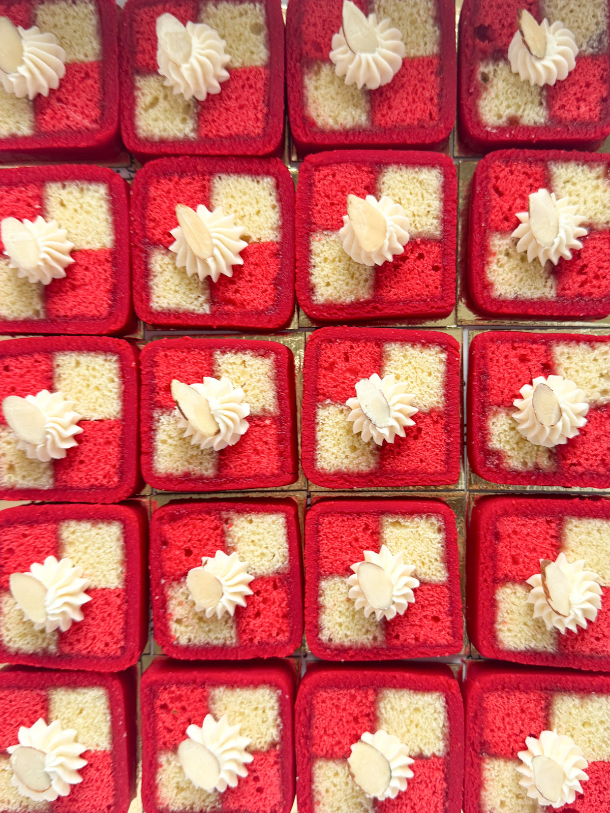 A grid of small square red and white layered cakes, known as battenberg cakes, each topped with a piped white cream swirl and a sliced almond on top, photographed from above.