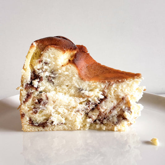 A slice of creamy burnt basque cheesecake with dark cinnamon swirls, with a golden-brown baked exterior, served on a white plate against a light gray background.