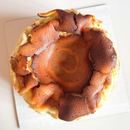 A top-down view of a cinnamon burnt basque cheesecake with a golden-brown spiral center and toasted around the edges. Placed on a white square board against a light background.