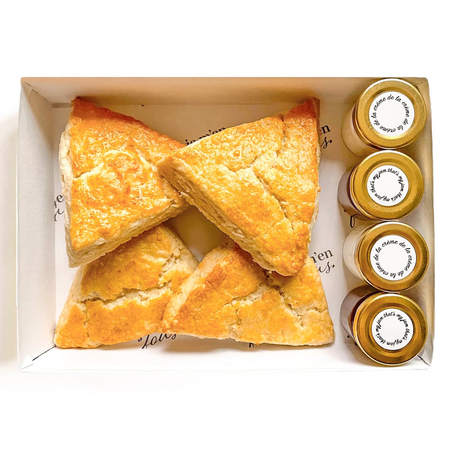 Box of triangle plain butter scones with jars of jam and clotted cream on a white background.