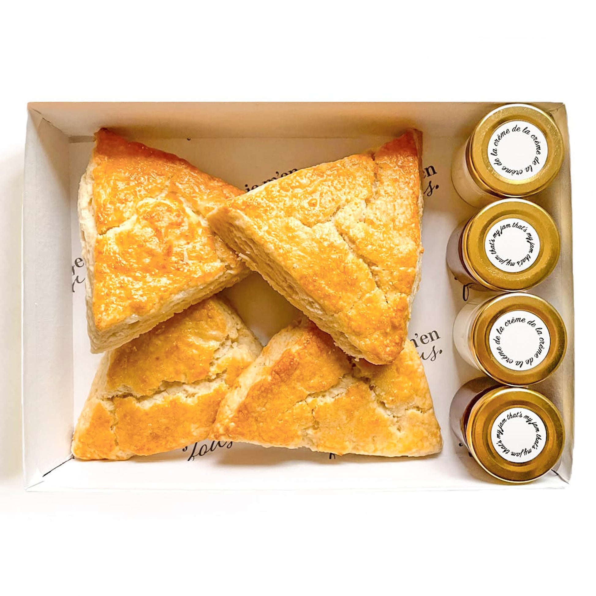 Box of triangle plain butter scones with jars of jam and clotted cream on a white background.