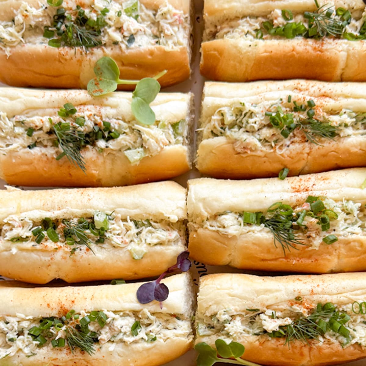 Close-up of multiple sliced sandwich rolls filled with shredded crab salad, topped with chopped green herbs and microgreens on a white background.