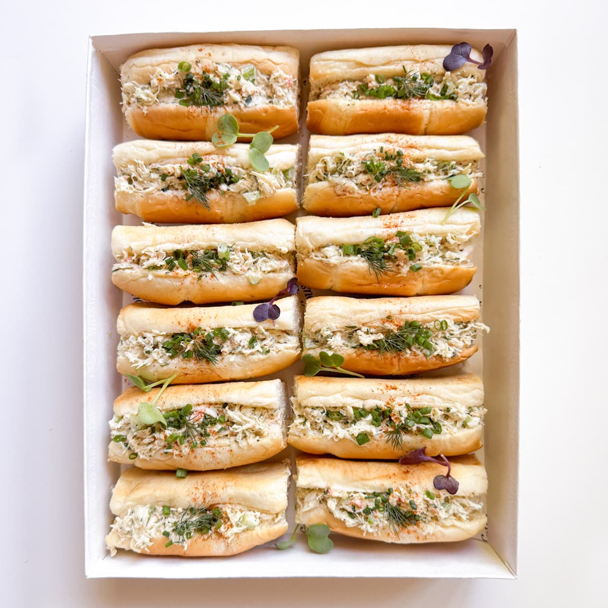 A top-down view of a white rectangular box containing twelve mini open-faced sandwiches filled with a creamy crab salad, garnished with microgreens.