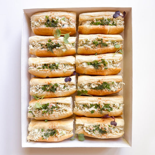 A top-down view of a white rectangular box containing twelve mini open-faced sandwiches filled with a creamy crab salad, garnished with microgreens.