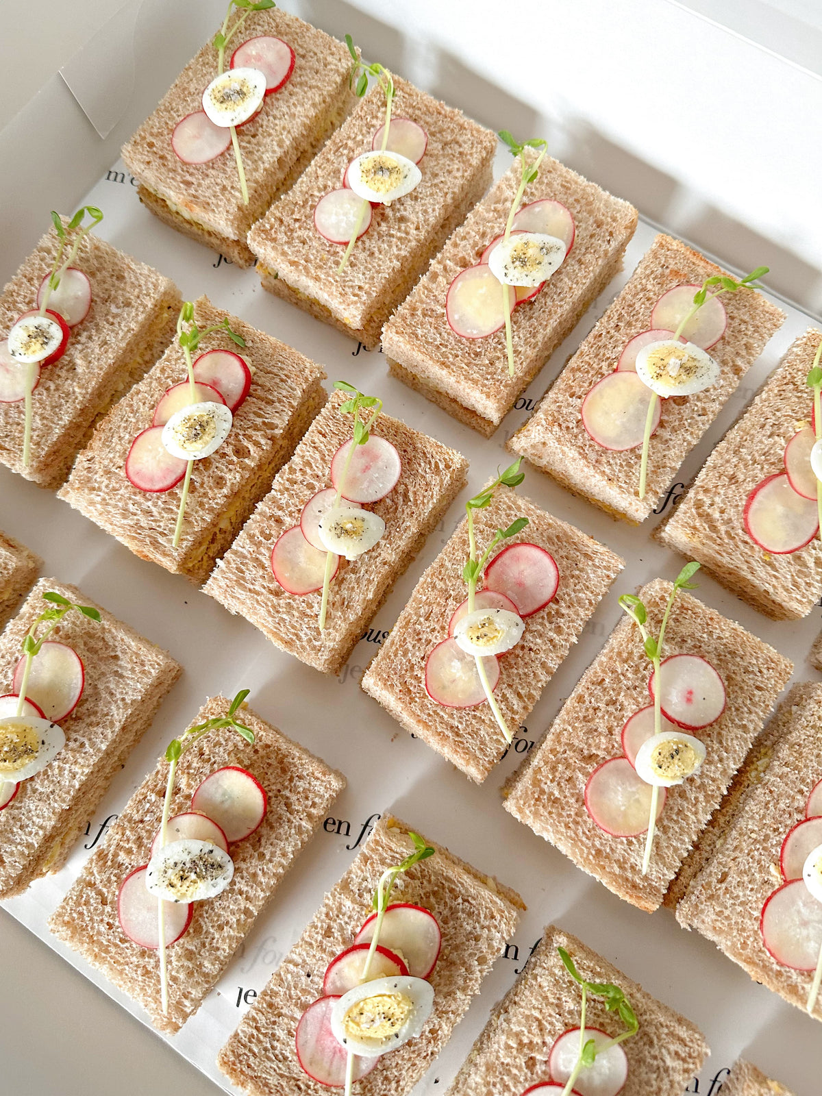 A top-down view of multiple small square sandwiches arranged in rows on a white tray. Each sandwich is made with brown bread and topped with sliced radishes, a sliced quail egg, and green sprouts.