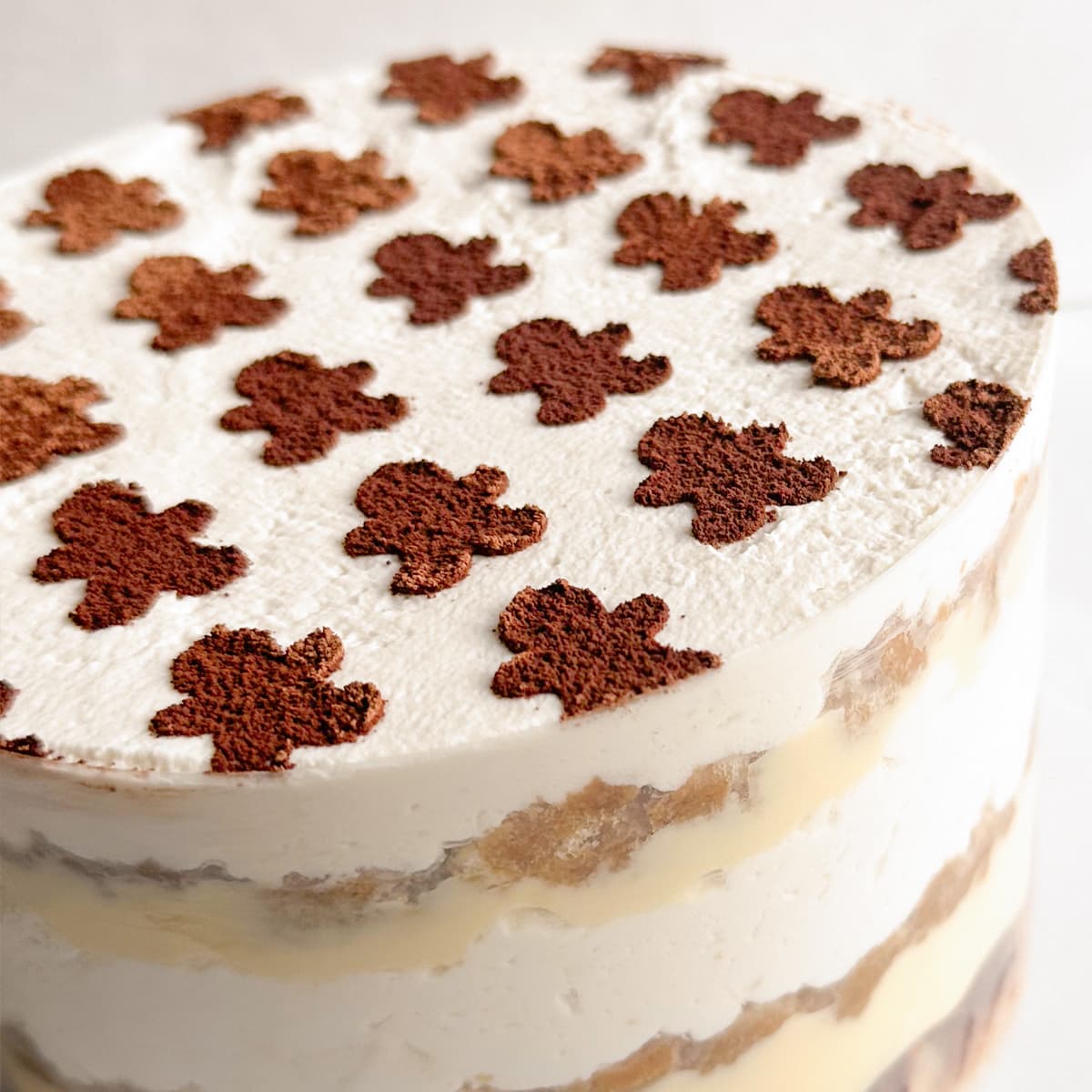 A close-up of a round white cake with alternating layers of cream and sponge cake, topped with small cocoa powder gingerbread-shaped decorations on a white background.