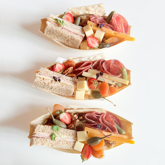 Three beautifully curated charcuterie boats arranged vertically on a clean white background, containing one ham & cheese tea sandwich, cheeses, cured meats, assorted nuts and seasonal fruit.