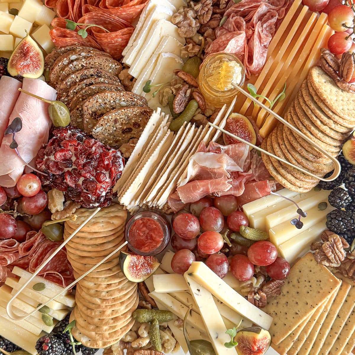 An overhead flat-lay of a charcuterie board with sliced cured meats, cheeses, crackers, nuts, grapes, figs, pickles, and small jars, arranged in a colorful food spread.