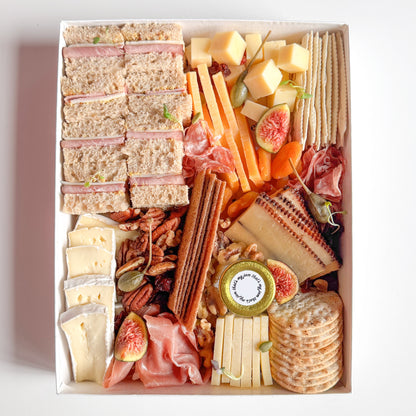 Assorted charcuterie board with meats, cheeses, mini tea sandwiches, crackers, and fruits in a box.