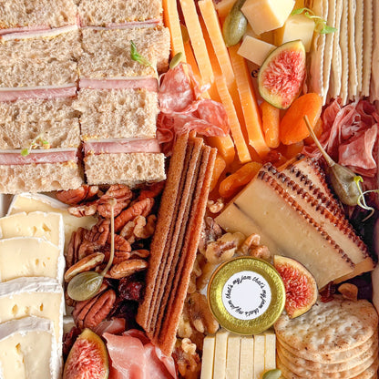 Assorted charcuterie board with meats, cheeses, mini tea sandwiches, fresh fruit, and crackers in a box, perfect for gifting.