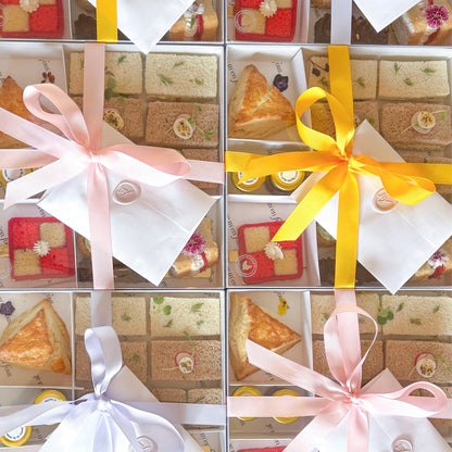 A grid of six gift boxes containing assorted baked goods, sandwiches, pastries, and small jars, each tied with a ribbon in a different pastel color: pink, yellow, and white. The boxes are arranged in a clean, symmetrical layout.