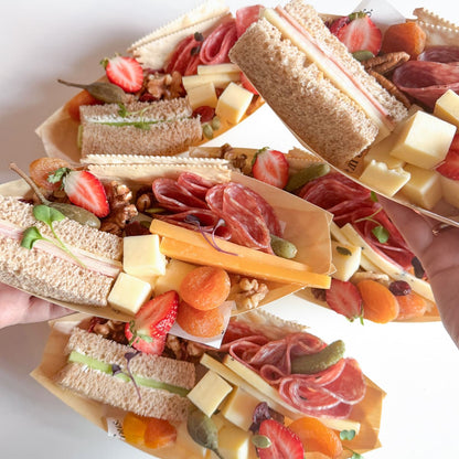 Several mini charcuterie sandwich boats filled with sliced bread, cheese cubes, strawberries, salami, prosciutto, nuts, and dried apricots are arranged on a white surface, with hands holding some of the boats.