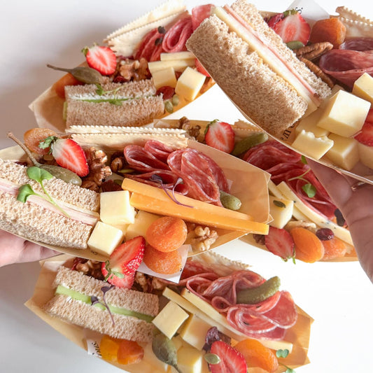 Several mini charcuterie sandwich boats filled with sliced bread, cheese cubes, strawberries, salami, prosciutto, nuts, and dried apricots are arranged on a white surface, with hands holding some of the boats.