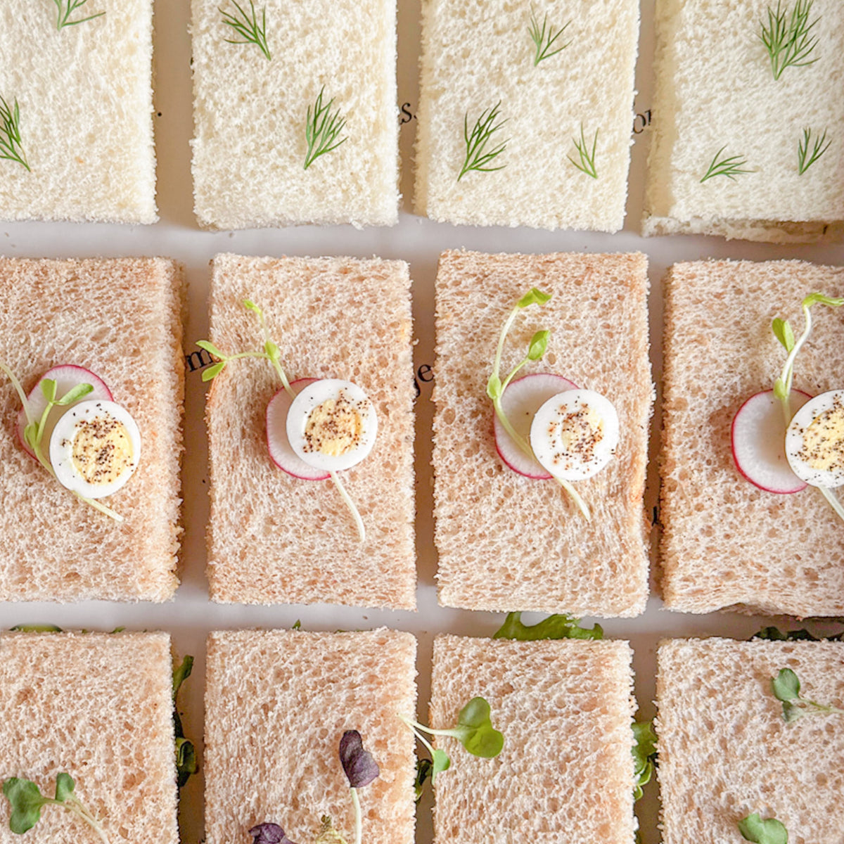 Overhead view of a grid of small tea sandwiches made with white and brown bread, topped with sliced hard-boiled eggs, radish slices, and microgreens.